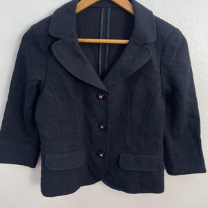 May include: A black blazer with three buttons and two front pockets. The blazer has three-quarter sleeves and a notched collar. The garment is hanging on a wooden hanger.