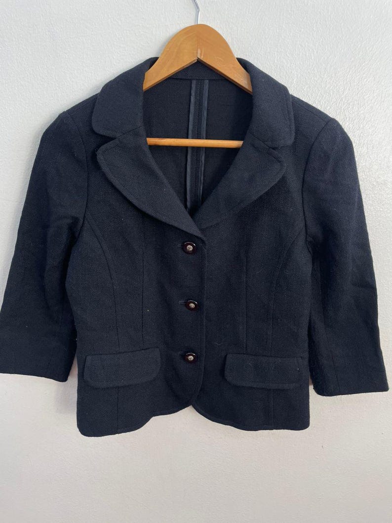 May include: A black blazer with three buttons and two front pockets. The blazer has three-quarter sleeves and a notched collar. The garment is hanging on a wooden hanger.