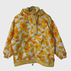 May include: A vintage anorak jacket with a floral pattern in yellow and orange on a white background. The jacket has a hood, a button-up front, and two front pockets with yellow zippers. The cuffs and hem are yellow.