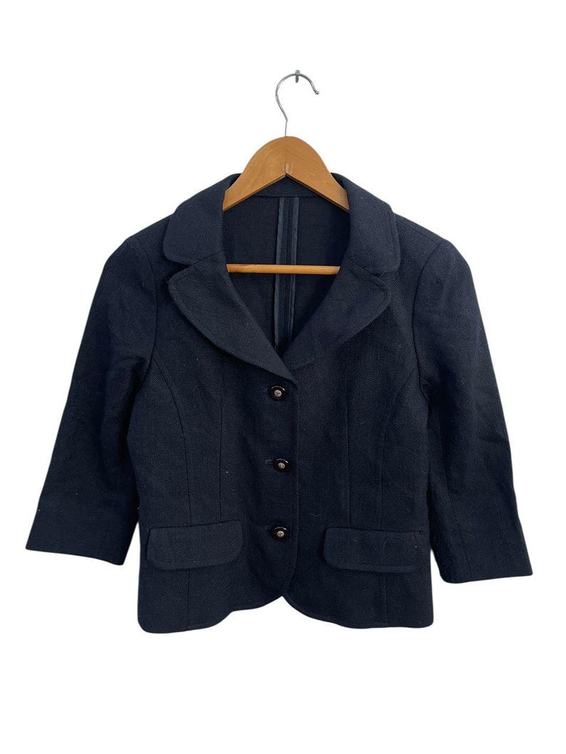 May include: A black blazer with three buttons and two front pockets, hanging on a wooden hanger. The blazer has three-quarter sleeves and a classic lapel design. The garment is made of a textured fabric.