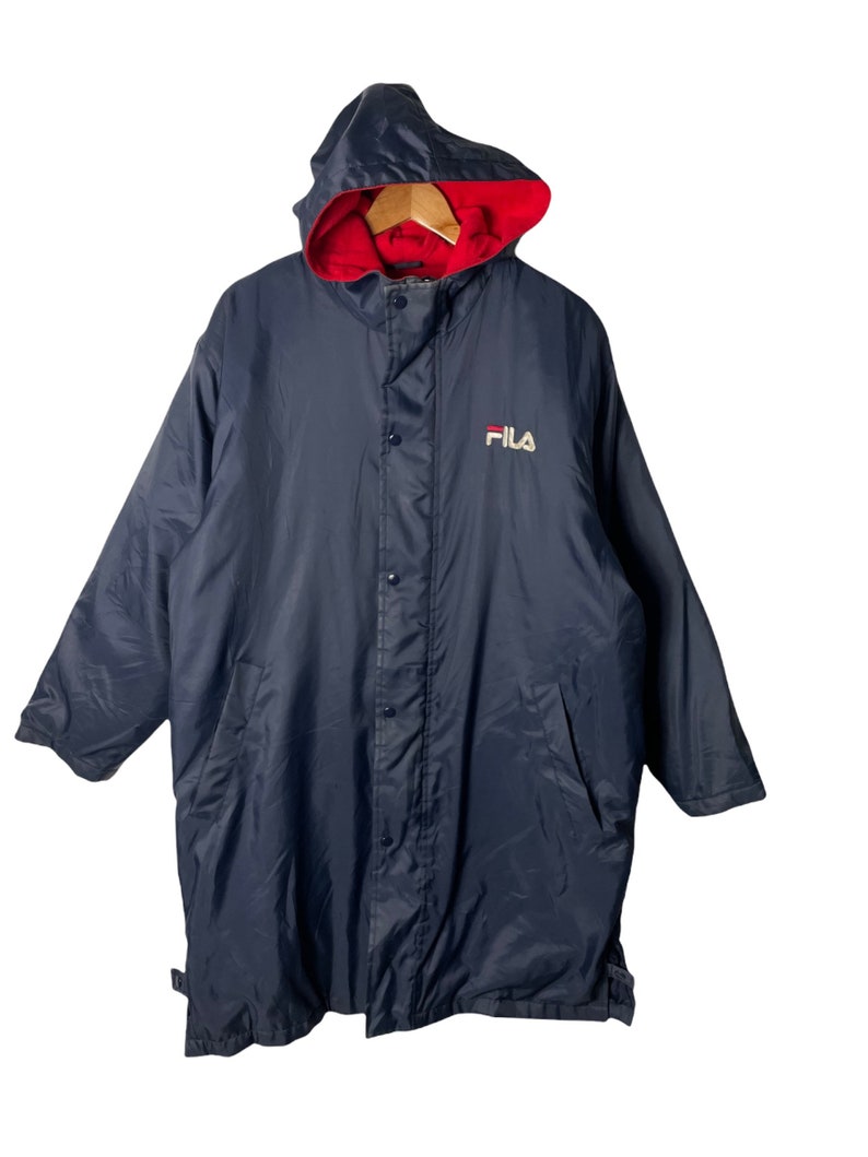 Pu&ograve; includere: Una giacca blu navy con cappuccio, fodera rossa e logo FILA bianco sul davanti. La giacca ha una chiusura con bottoni e due tasche.