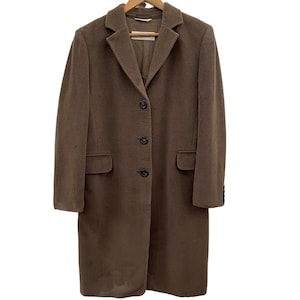 Puede incluir: Un abrigo largo de manga larga de color marrón con cierre de botones y dos bolsillos. El abrigo está colgado en una percha de madera.