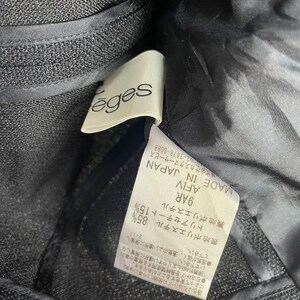 May include: Close-up of a dark gray fabric with a label attached. The label reads "&eacute;ges" in a cursive font. Additional text on the label includes Japanese characters and fabric composition details. The fabric appears to be a textured weave.
