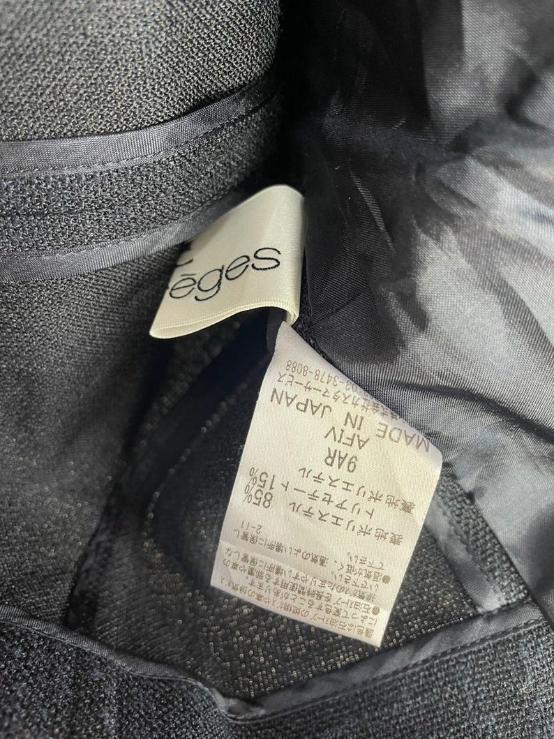 May include: Close-up of a dark gray fabric with a label attached. The label reads "&eacute;ges" in a cursive font. Additional text on the label includes Japanese characters and fabric composition details. The fabric appears to be a textured weave.
