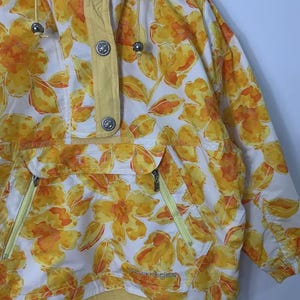 May include: A vintage anorak jacket featuring a vibrant floral pattern in shades of yellow and orange on a white background. The jacket has a hood, a front pocket with zippers, and a yellow trim. The jacket has silver buttons and drawstrings.