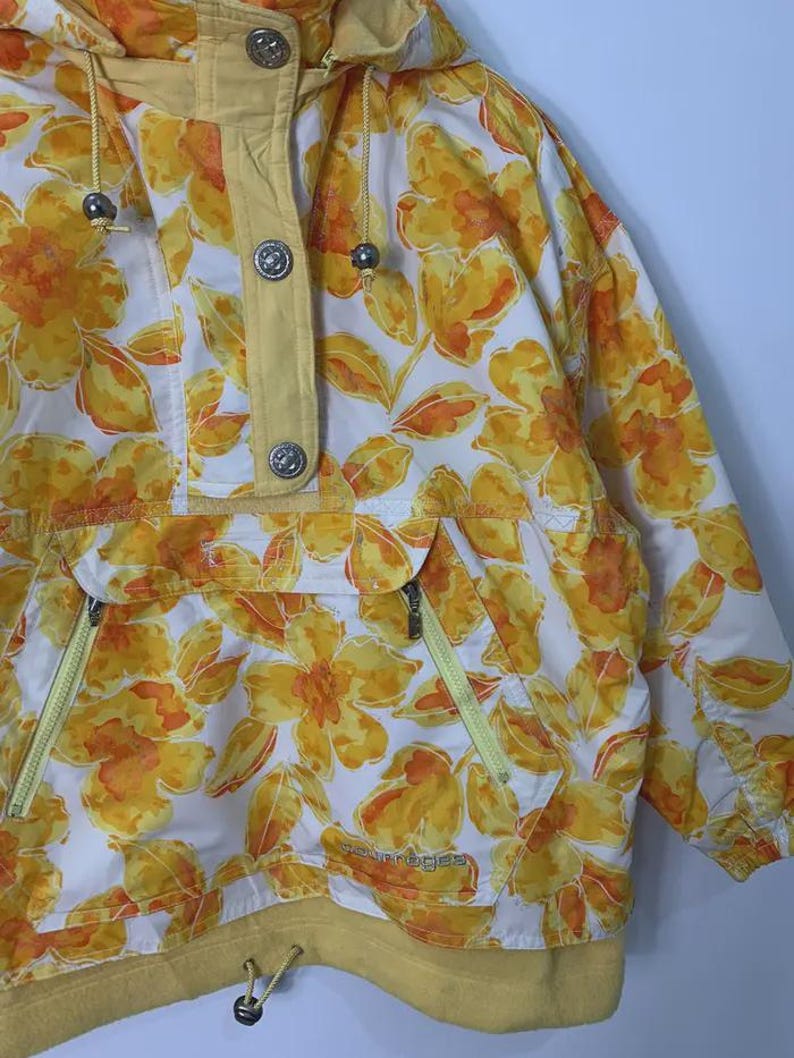 May include: A vintage anorak jacket featuring a vibrant floral pattern in shades of yellow and orange on a white background. The jacket has a hood, a front pocket with zippers, and a yellow trim. The jacket has silver buttons and drawstrings.