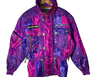Puede incluir: Una chaqueta de esquí morada y rosa con un patrón geométrico. La chaqueta tiene cierre de cremallera, varios bolsillos y cintura con cordón. El nombre de la marca "SALOMON" está impreso en la parte delantera de la chaqueta.