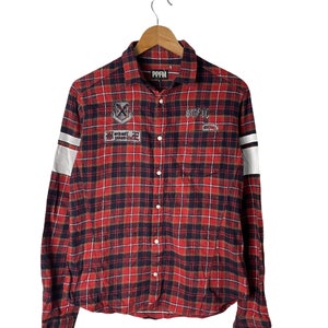 Puede incluir: Camisa de manga larga a cuadros rojos y negros con botones, con rayas blancas en las mangas y varios parches, incluyendo "PPFM" y "WORK HARD PLAY HARD".