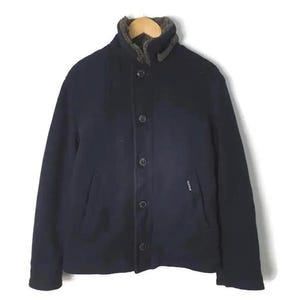 Puede incluir: Chaqueta azul marino con botones y cuello de piel sintética. La chaqueta tiene un diseño clásico con una estética sencilla y limpia. La chaqueta está colgada en una percha de madera.