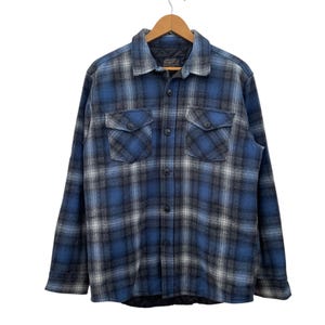 May include: Blue, black, and white plaid flannel shirt with two chest pockets and button closures. The long-sleeved shirt is hanging on a wooden hanger. The shirt has a quilted lining.