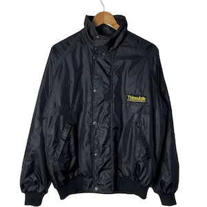 Puede incluir: Una chaqueta de nylon negra con cuello alto y cierre de presión. La chaqueta tiene un logotipo de Thinsulate en el pecho izquierdo.