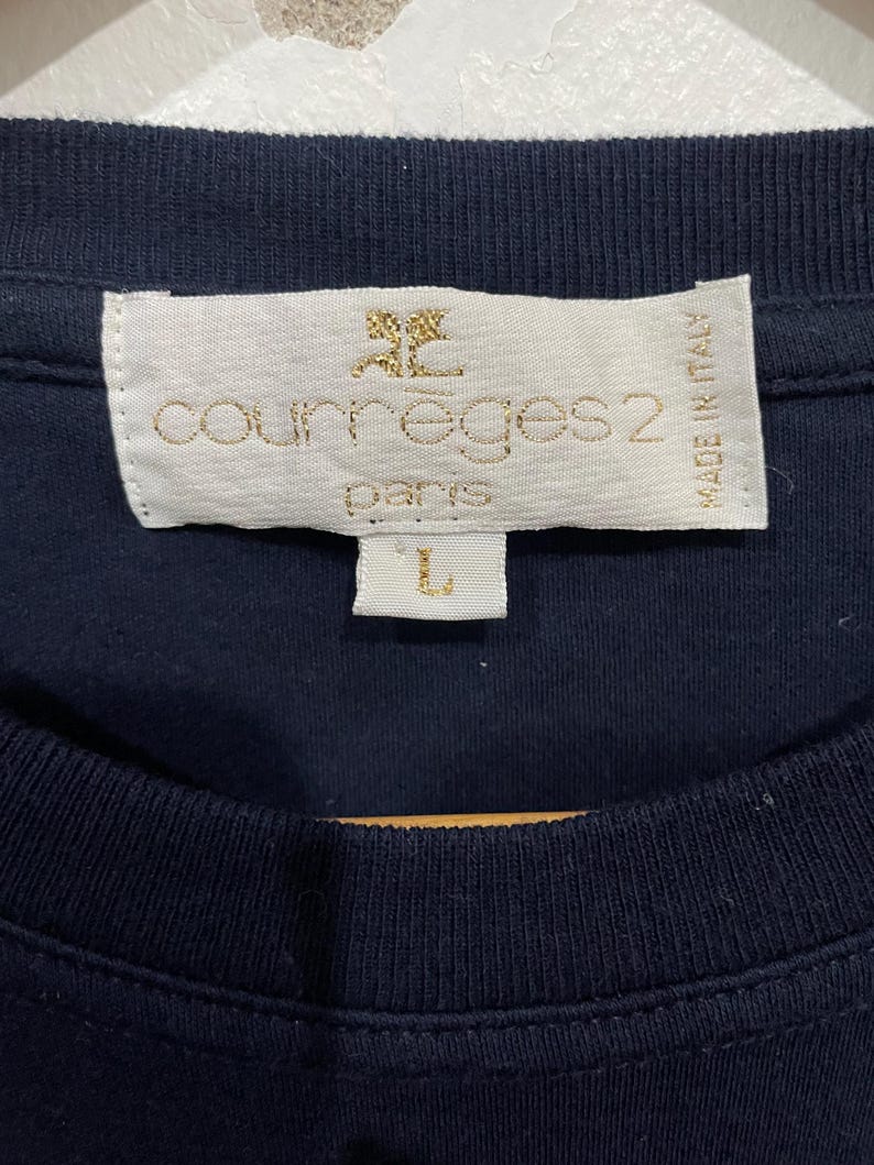 Op de afbeelding: Een marineblauw T-shirt met een wit label waarop "Courr&egrave;ges 2 Paris" en "Made in Italy" staat. Het label heeft ook een klein gouden logo van een gestileerde "C".