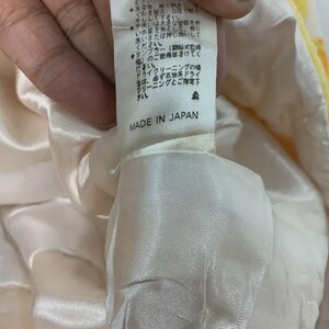 May include: A close-up of a tag with Japanese text and the words "MADE IN JAPAN" printed in black. The tag is attached to a cream-colored fabric with a yellow floral pattern.