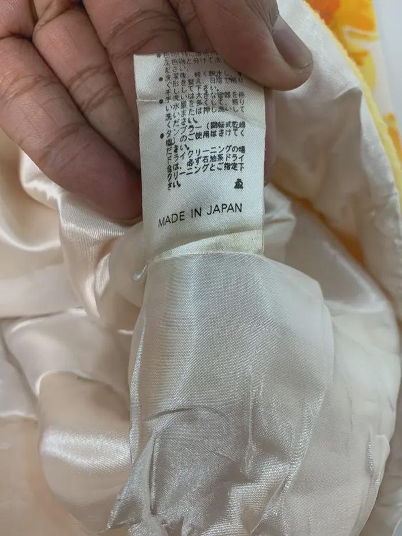 May include: A close-up of a tag with Japanese text and the words "MADE IN JAPAN" printed in black. The tag is attached to a cream-colored fabric with a yellow floral pattern.