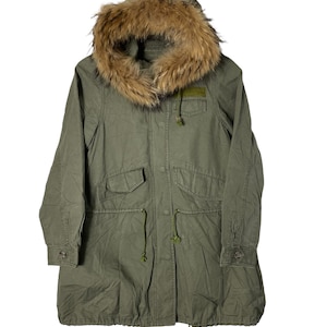 Puede incluir: Una parka verde con capucha y un ribete de piel sintética marrón alrededor de la capucha. La chaqueta tiene una cintura con cordón y varios bolsillos.