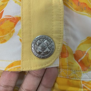 May include: Close-up of a section of a garment with a yellow and floral pattern. A silver button with the text "Courreges Sports" is visible. The garment has a yellow trim and a white background.