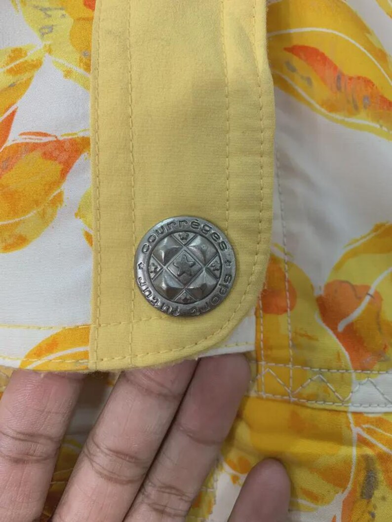 May include: Close-up of a section of a garment with a yellow and floral pattern. A silver button with the text "Courreges Sports" is visible. The garment has a yellow trim and a white background.
