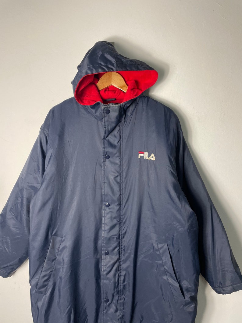 Pu&ograve; includere: Una giacca imbottita con cappuccio blu navy con fodera rossa e un logo FILA bianco sul davanti.