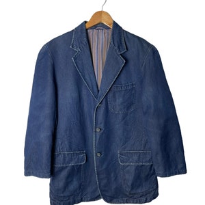 Puede incluir: Una chaqueta de sport de mezclilla azul oscuro con cierre de botones y varios bolsillos. La chaqueta está colgada en una percha de madera.
