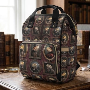 May include: A structured backpack featuring a repeating pattern of framed portraits of bearded dragons dressed in formal Victorian-style attire. The bag has black top handles, a front zippered pocket, and gold-toned hardware accents.