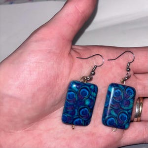 May include: A pair of rectangular earrings with a peacock feather design. The earrings feature a blue and teal pattern with silver-colored hooks. The earrings are approximately 1 inch long and 0.75 inches wide.