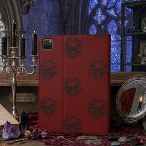 Peut inclure: Une housse de tablette rouge foncé ornée d'un motif répétitif noir représentant des crânes entourés de roses. L'étui rectangulaire présente une finition mate lisse et une découpe pour l'appareil photo dans le coin supérieur gauche.
