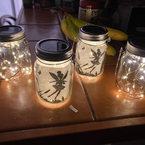 Puede incluir: Cuatro tarros de cristal tipo Mason con tapas negras y asas de alambre. Dos tarros presentan siluetas de hadas sobre papel, iluminadas por luces interiores. Los otros dos tarros están llenos de luces de cadena. Los tarros están colocados sobre una superficie marrón.