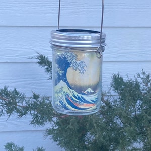 May include: A clear glass mason jar lantern featuring a wrap-around print of a large blue ocean wave and a mountain. The jar has a silver-colored metal lid and a thin, arched wire handle attached to the sides for hanging purposes.
