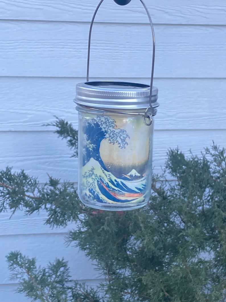 May include: A clear glass mason jar lantern featuring a wrap-around print of a large blue ocean wave and a mountain. The jar has a silver-colored metal lid and a thin, arched wire handle attached to the sides for hanging purposes.