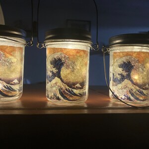May include: A set of clear glass mason jars with black lids and wire handles, each illuminated from within. The exterior of each jar features a printed graphic of a large blue ocean wave with white foam crests and a mountain in the background.