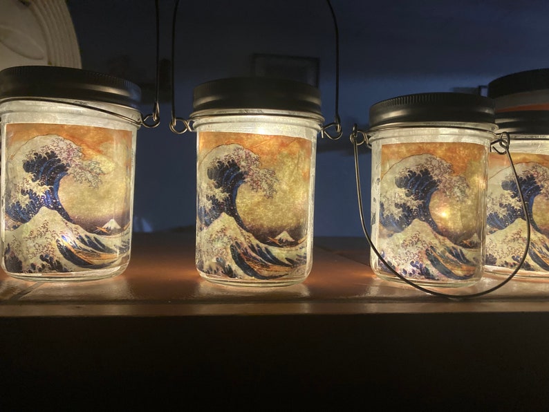May include: A set of clear glass mason jars with black lids and wire handles, each illuminated from within. The exterior of each jar features a printed graphic of a large blue ocean wave with white foam crests and a mountain in the background.
