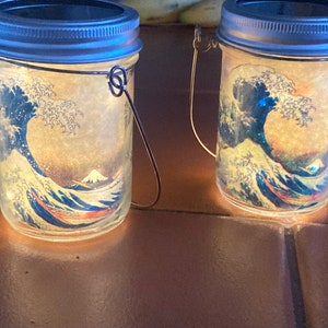 Può includere: Due barattoli di vetro con una luce solare al loro interno. I barattoli hanno un manico in metallo e un corpo in vetro trasparente. I barattoli sono decorati con una stampa della Grande Onda di Kanagawa di Hokusai.
