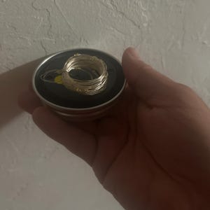 Puede incluir: Una tapa metálica circular de color plateado que presenta una hebra enrollada de alambre de cobre fino y flexible con pequeñas luces LED integradas. La tapa tiene un inserto central negro plano y un borde metálico brillante.