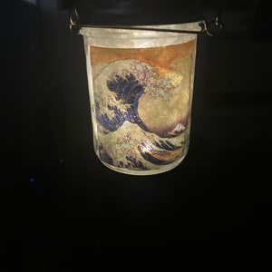 May include: A glass jar lantern featuring a wrap-around print of The Great Wave off Kanagawa. The jar has a dark metal lid with a wire handle for hanging. The interior is illuminated, casting a warm glow through the translucent artwork.