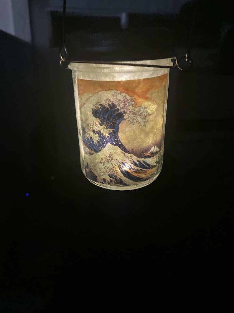 May include: A glass jar lantern featuring a wrap-around print of The Great Wave off Kanagawa. The jar has a dark metal lid with a wire handle for hanging. The interior is illuminated, casting a warm glow through the translucent artwork.