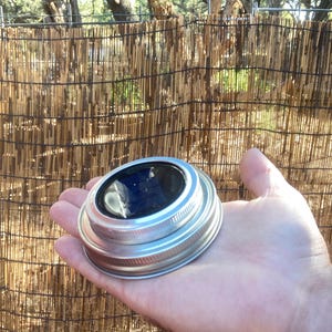 May include: A solar-powered mason jar lid with a silver metal rim and a dark blue solar panel. The lid is held in a hand, with a natural bamboo fence in the background. The solar panel is designed to charge during the day.