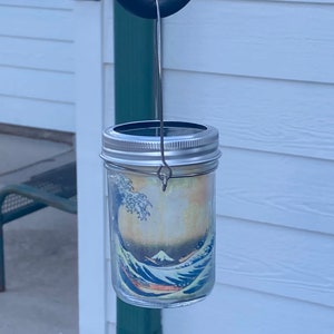 May include: A clear glass mason jar lantern featuring a wrap-around print of The Great Wave off Kanagawa. The jar has a silver-colored metal lid with a solar panel top and a thin wire handle attached to the neck for hanging purposes.