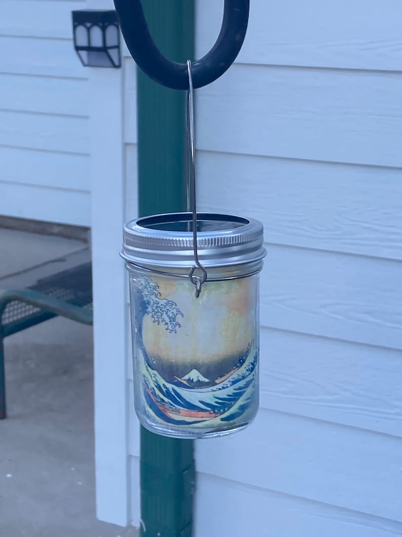 May include: A clear glass mason jar lantern featuring a wrap-around print of The Great Wave off Kanagawa. The jar has a silver-colored metal lid with a solar panel top and a thin wire handle attached to the neck for hanging purposes.