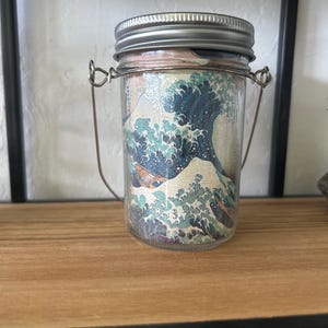 May include: A clear glass jar with a silver metal screw-top lid and a thin wire handle. The exterior of the jar features a wrap-around print of a large blue ocean wave with white foam crests, reminiscent of a classic Japanese woodblock art style.