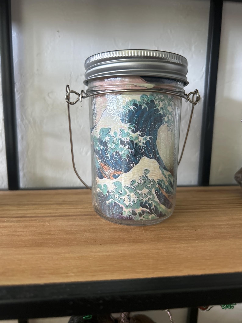 May include: A clear glass jar with a silver metal screw-top lid and a thin wire handle. The exterior of the jar features a wrap-around print of a large blue ocean wave with white foam crests, reminiscent of a classic Japanese woodblock art style.