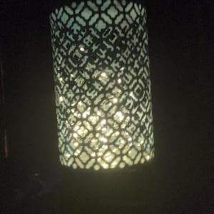 Puede incluir: Una linterna cilíndrica decorativa con un diseño calado. El material oscuro presenta un patrón recortado que permite el paso de la luz. El interior está iluminado con un brillo cálido, creando un efecto decorativo.