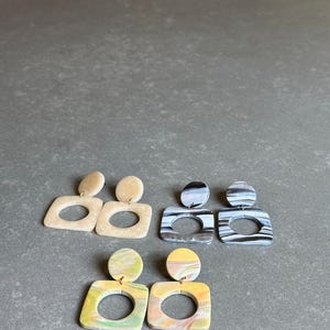 May include: Four pairs of square-shaped earrings with a circle cutout. The earrings are made of marbled resin in beige, black and white, and yellow and green.