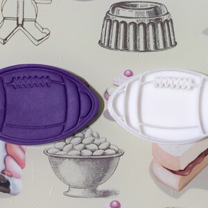 Può includere: Due formine per biscotti a forma di pallone da football. Uno è viola pieno, l'altro bianco. Entrambi hanno cuciture e lacci dettagliati. Le formine sono su una superficie a motivi con illustrazioni di dolci.