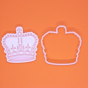 May include: Two white plastic crown-shaped cookie cutters. One is a detailed crown with decorative elements, the other a simple outline. The cutters are on an orange background.