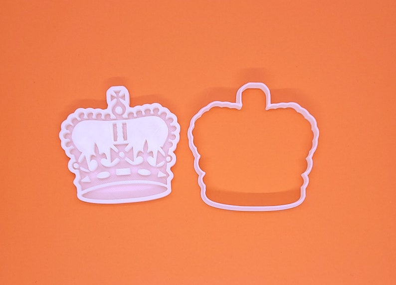 May include: Two white plastic crown-shaped cookie cutters. One is a detailed crown with decorative elements, the other a simple outline. The cutters are on an orange background.