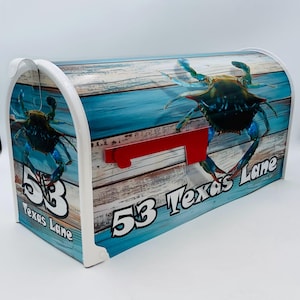 May include: A white mailbox with a blue crab design and the text "53 Texas Lane." The mailbox has a red flag and a wooden plank background with blue accents. The crab is blue and black.