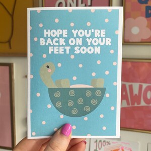 May include: A blue and white card with pink polka dots. The card features a cartoon turtle in a bowl and the text "Hope you're back on your feet soon."