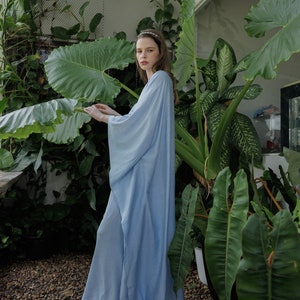 K&ouml;nnte beinhalten: Eine Frau in einem langen, hellblauen, flie&szlig;enden Kleid mit langen &Auml;rmeln steht in einer Gartenlandschaft. Sie ist umgeben von &uuml;ppigen gr&uuml;nen Pflanzen mit gro&szlig;en Bl&auml;ttern.