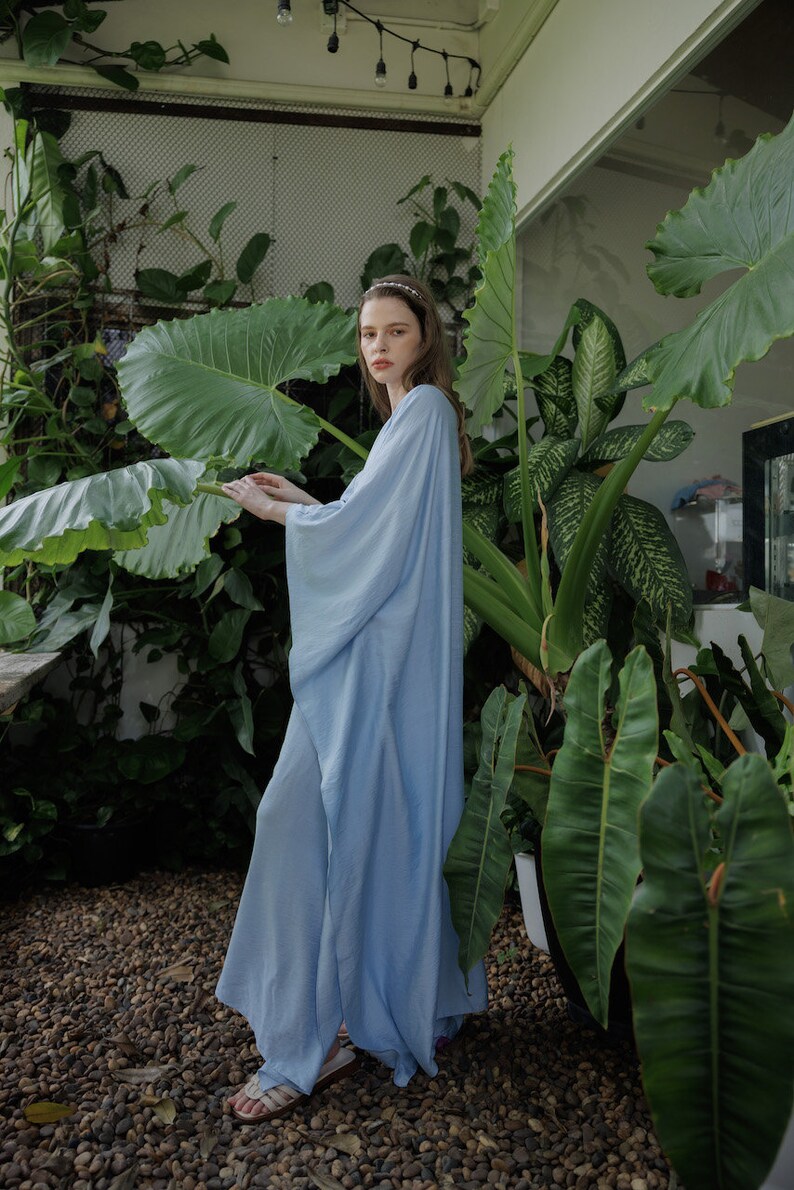 K&ouml;nnte beinhalten: Eine Frau in einem langen, hellblauen, flie&szlig;enden Kleid mit langen &Auml;rmeln steht in einer Gartenlandschaft. Sie ist umgeben von &uuml;ppigen gr&uuml;nen Pflanzen mit gro&szlig;en Bl&auml;ttern.
