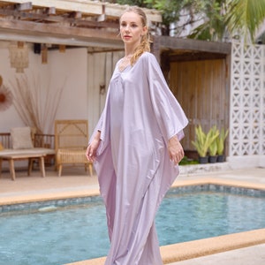 Puede incluir: Una mujer con un vestido largo de caft&aacute;n de color lila claro y fluido. Est&aacute; de pie junto a una piscina y mirando hacia la izquierda del encuadre.